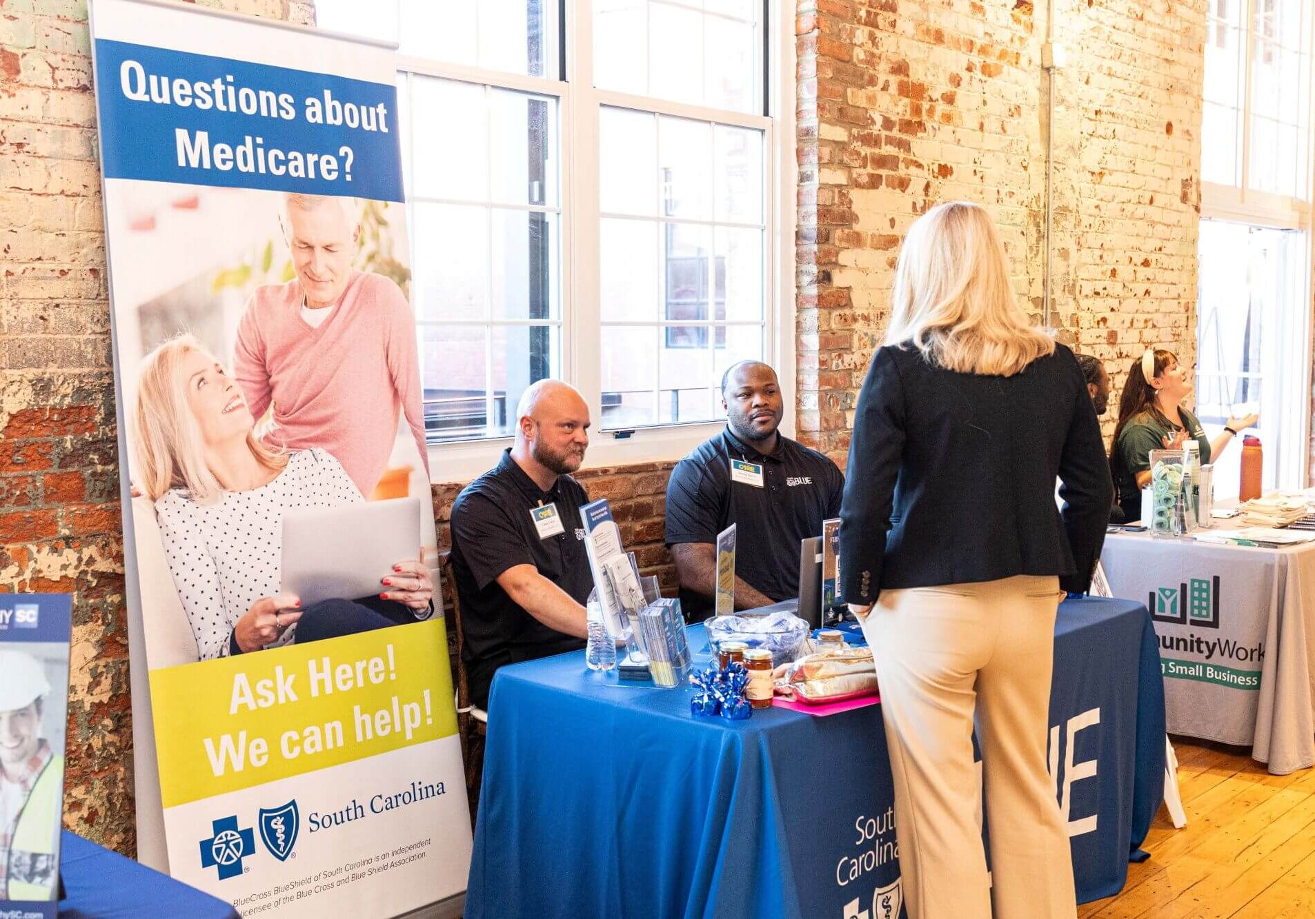 Exhibitors at a table with a sign reading "Questions about Medicare? Ask Here! We can help"